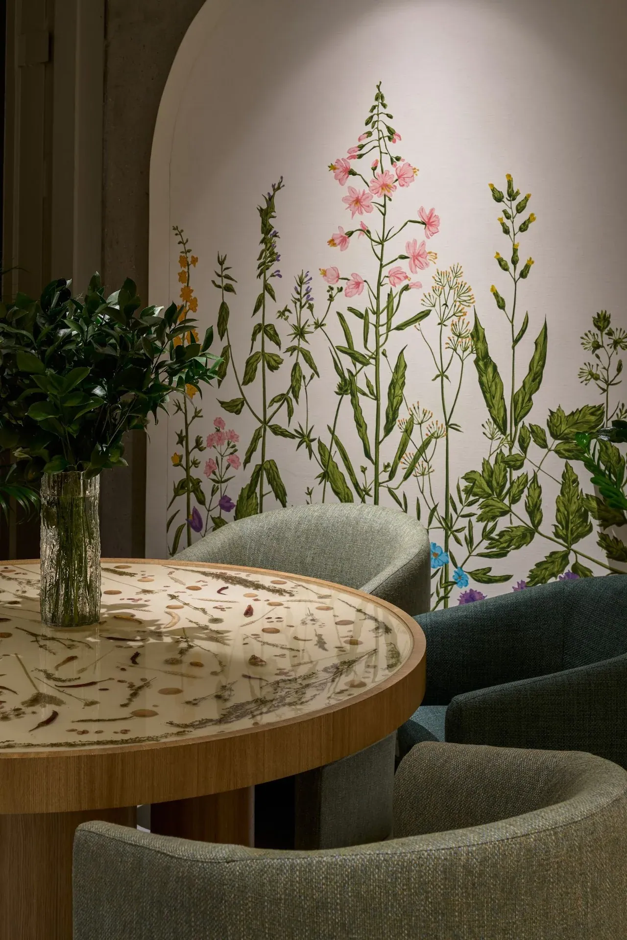 Round table with a botanical top in front of a painted botanical wall, with green armchairs