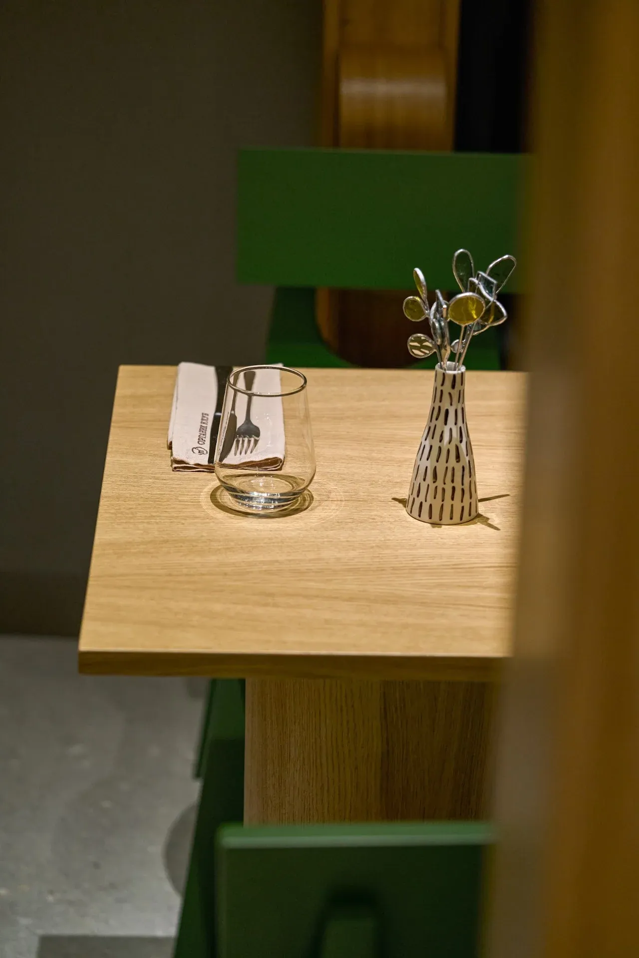 Oak table set for two with a small vase, beside oak pylons and green chairs