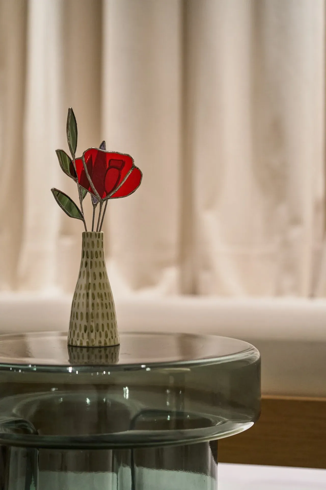 A red rose in a ceramic vase on a glass table in front of a cream curtain